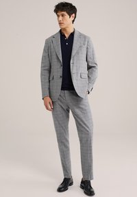 Sivý kockovaný oblek s klopovými reverzami, v kombinácii s námorníckym polo tričkom. Čierne topánky dopĺňajú outfit. Prispôsobený strih a hladká textúra.