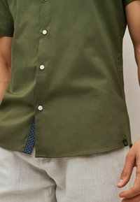 La camisa de manga corta en color oliva presenta botones blancos, una abertura lateral con un interior de patrón marino y una tela con textura sutil.
