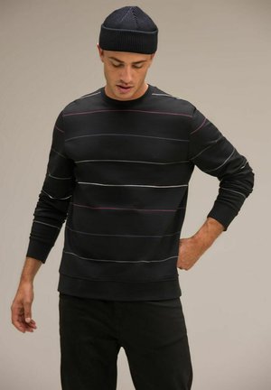 Sweatshirt noir avec des rayures horizontales en blanc, rose et bleu, poignets côtelés et col rond. Porté avec un pantalon noir et un bonnet en tricot.