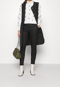 Czarna pikowana kamizelka bez rękawów na białym swetrze w czarne kropki, do tego czarne obcisłe spodnie i kremowe botki, trzymająca oliwkową torbę.