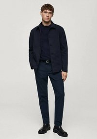 Giacca in misto lana blu navy con chiusura a bottoni, abbinata a un maglione a collo rotondo blu scuro e pantaloni su misura. Stivali in pelle nera completano il look.