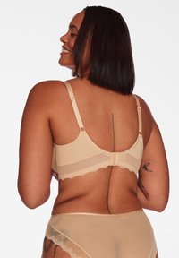 Kobieta o średnim odcieniu skóry i prostych czarnych włosach uśmiecha się, ubrana w beżową bieliznę z widocznymi tatuażami na plecach i ramionach, odwrócona plecami.