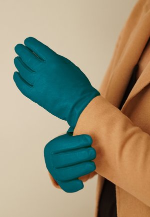 Teal leather gloves with a smooth texture, featuring a fitted design, finger seams, and a snug cuff, worn on a beige coat.