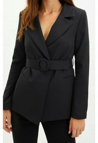 Musta räätälöity bleiseri, jossa on vyötärövyö, syvä V-aukko, pitkät hihat ja kaksi sivutaskua. Sileä kangas ja jäsennelty muotoilu.