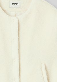 Veste en polaire blanche avec un col rond, fermeture à boutons pression et poches latérales. Tissu doux et texturé, design minimaliste. Étiquette apposée.