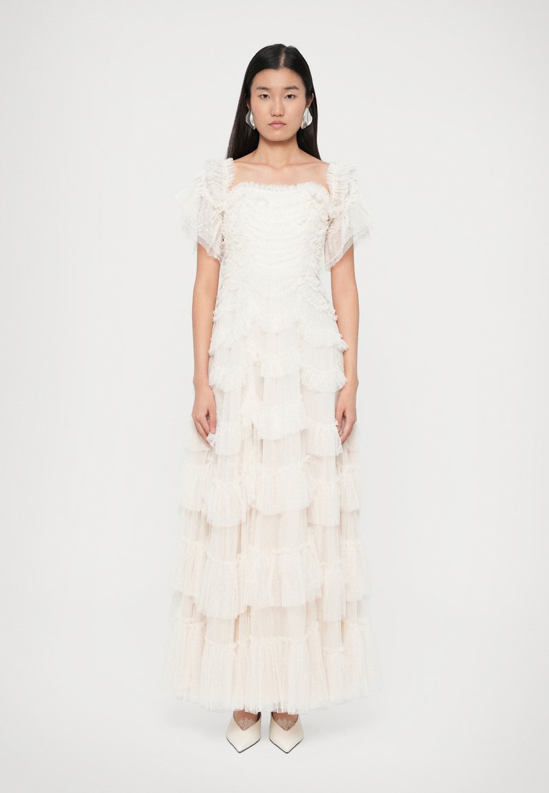 Bílé tiered šaty z tylu s volánkovými vrstvami a krátkými nabíranými rukávy. Modelka má na sobě bílé lodičky s špičatou špičkou.