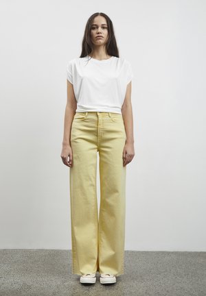 T-shirt bianco a maniche corte abbinato a pantaloni gialli a vita alta e gamba larga. Tessuto morbido con una vestibilità rilassata. Modello che indossa sneakers bianche.