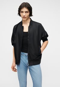 Νεαρός με μεσαίου μήκους σκούρα μαλλιά, φοράει μαύρο μπουφάν με φερμουάρ πάνω από μαύρο τοπ και ανοιχτό μπλε τζιν, με τα χέρια στις τσέπες.