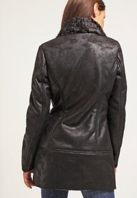 Abrigo de cuero sintético negro con un acabado texturizado, que cuenta con un collar de piel, una silueta ajustada y detalles de costura en la parte posterior.