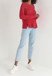 Punainen resorineulottu villapaita sivupalmikoilla, yhdistetty vaaleansinisiin suoran lahkeen farkkuihin ja valkoisiin paksupohjaisiin lenkkareihin.