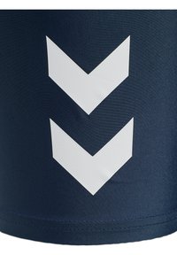 Två vita nedåtriktade chevronpilar på mörkblått texturerat tyg, med synliga stygn vid nedre kanten.