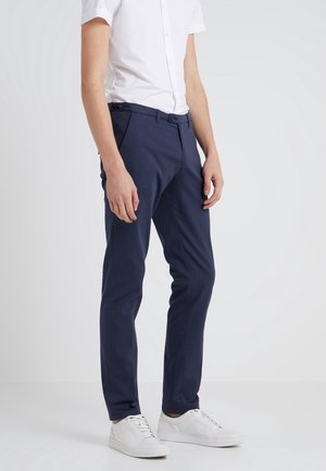 Mężczyzna ubrany w granatowe, dopasowane spodnie, białą koszulę z krótkim rękawem na guziki oraz białe trampki, stojący na jednolitym jasnym tle.