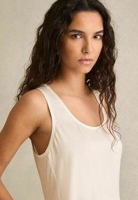 Giovane donna con lunghi capelli ricci scuri che indossa una canotta crema, in piedi davanti a uno sfondo beige chiaro uniforme.