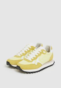 Gula träningsskor med ovandel i mocka, vita detaljer och en texturerad svart gummisula. Har traditionell snörning och vadderad krage.