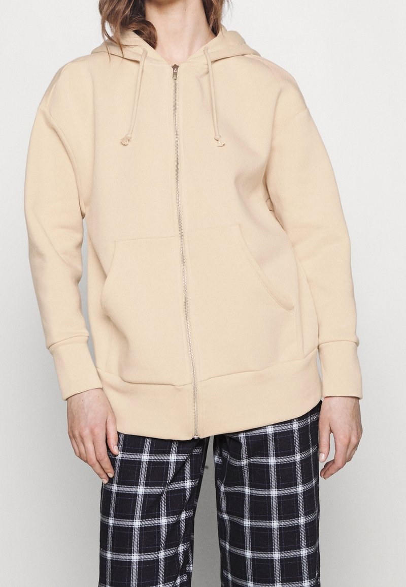 Sweat à capuche beige fermé par une fermeture éclair, fabriqué en matière douce, doté d'une poche kangourou devant et d'une capuche avec cordon de serrage. Associé à un pantalon noir et blanc à carreaux.