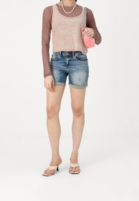Haut sans manches rose clair en tissu texturé, superposé sur une chemise marron à manches longues. Porté avec un short en jean et des sandales à talons beiges.
