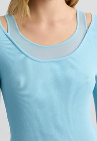 Haut ajusté bleu clair avec texture côtelée, empiècement transparent au niveau du décolleté et détails découpés aux épaules. Tissu lisse avec une coupe près du corps.