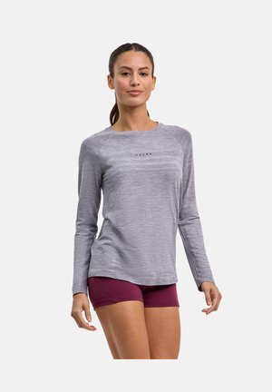 Kobieta o ciemnych włosach związanych w kucyk, ubrana w szarą sportową bluzę z długim rękawem i bordowe krótkie spodenki sportowe, stojąca na jednolitym tle.