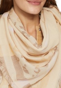 Beige-Schal mit weicher Textur und subtilen Tiermustern, mit fransigen Kanten und lässig um den Hals gewickelt.