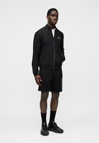 Melnā rāvējslēdzēja jaka ar tekstūras detaļām, kombinēta ar melniem šortiem. Melni sporta apavi un zeķes papildina sportisko apģērbu. Ietver logotipu akcentus.