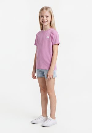 Uśmiechnięta dziewczyna stojąca, ubrana w różowy t-shirt z krótkim rękawem, jasne szorty jeansowe i białe trampki na jednolitym białym tle.