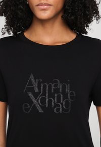 Čierne bavlnené tričko s potlačou zo strieborného trblietavého loga, na ktorom je nápis "Armani Exchange". Kolo výstrih a krátke rukávy.