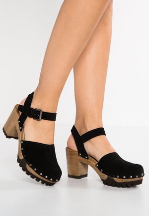 Czarne zamszowe klompy na obcasie z regulowanym paskiem na kostce, drewnianym obcasem, srebrnymi ćwiekami wzdłuż krawędzi oraz teksturowaną gumową podeszwą.