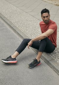 Schwarze Sportschuhe mit roten Akzenten, schwarze schmal geschnittene Hose und ein rotes kurzärmliges Hemd. Modell sitzt auf einem Kopfsteinpflaster-Bordstein.