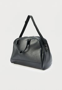 Sac de voyage en cuir noir avec une texture lisse, une forme incurvée, deux poignées et une bandoulière amovible. Présente des accents de quincaillerie argentée.