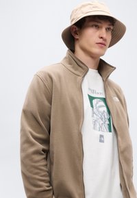 Μπεζ φλις μπουφάν με ψηλό γιακά και κλείσιμο με φερμουάρ, φορεμένο πάνω από λευκό μπλουζάκι με γραφικό σχέδιο, σε συνδυασμό με ανοιχτό μπεζ καπέλο τύπου bucket.