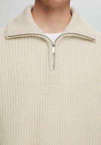 Jersey beige con cuello con media cremallera. Presenta un patrón texturizado y un cuello en gris claro contrastante, ofreciendo un aspecto acogedor y estructurado.