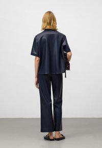 Osoba z blond włosami w czarnej koszulce z krótkim rękawem i odpowiadających jej szerokich spodniach, trzymająca ciemną torbę, stojąca na szarym podłodze przy białej ścianie.