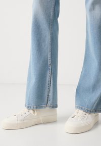 Vaqueros de mezclilla de color azul claro con un detalle en la costura lateral, combinados con zapatillas de lona blancas que tienen una suela texturizada y cordones planos.