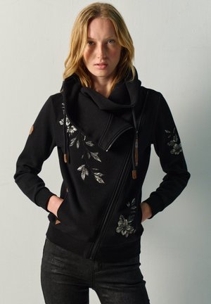 Mujer con cabello rubio que lleva una chaqueta negra con cremallera, bordado floral plateado y las manos en los bolsillos contra un fondo liso.