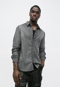 Tânărul poartă o cămașă gri cu nasturi peste un top negru și pantaloni negri din piele cargo, stând cu spatele la un fundal alb simplu.