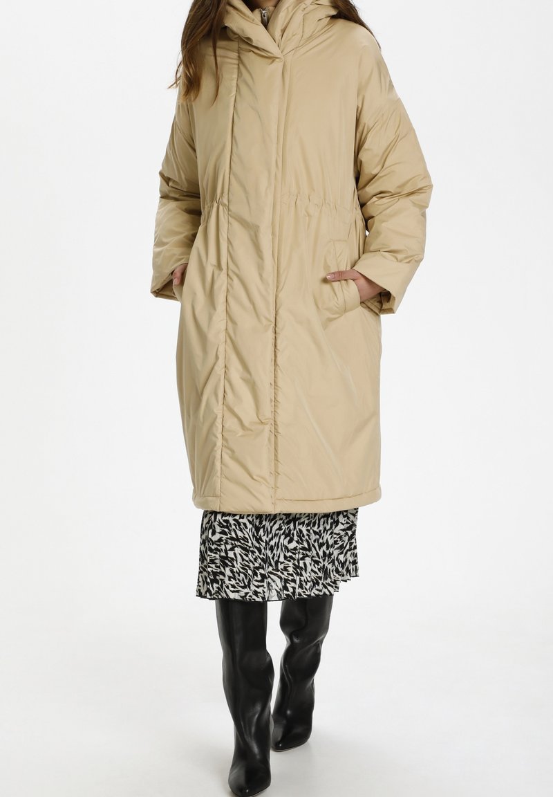 Femme portant un manteau matelassé beige par-dessus une jupe à motifs noir et blanc et des bottes noires montantes, debout les mains dans les poches du manteau.