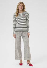 Top a maniche lunghe a righe bianco e nero con un piccolo dettaglio a forma di cuore rosso, abbinato a jeans ampi grigi chiari e scarpe col tacco rosse.