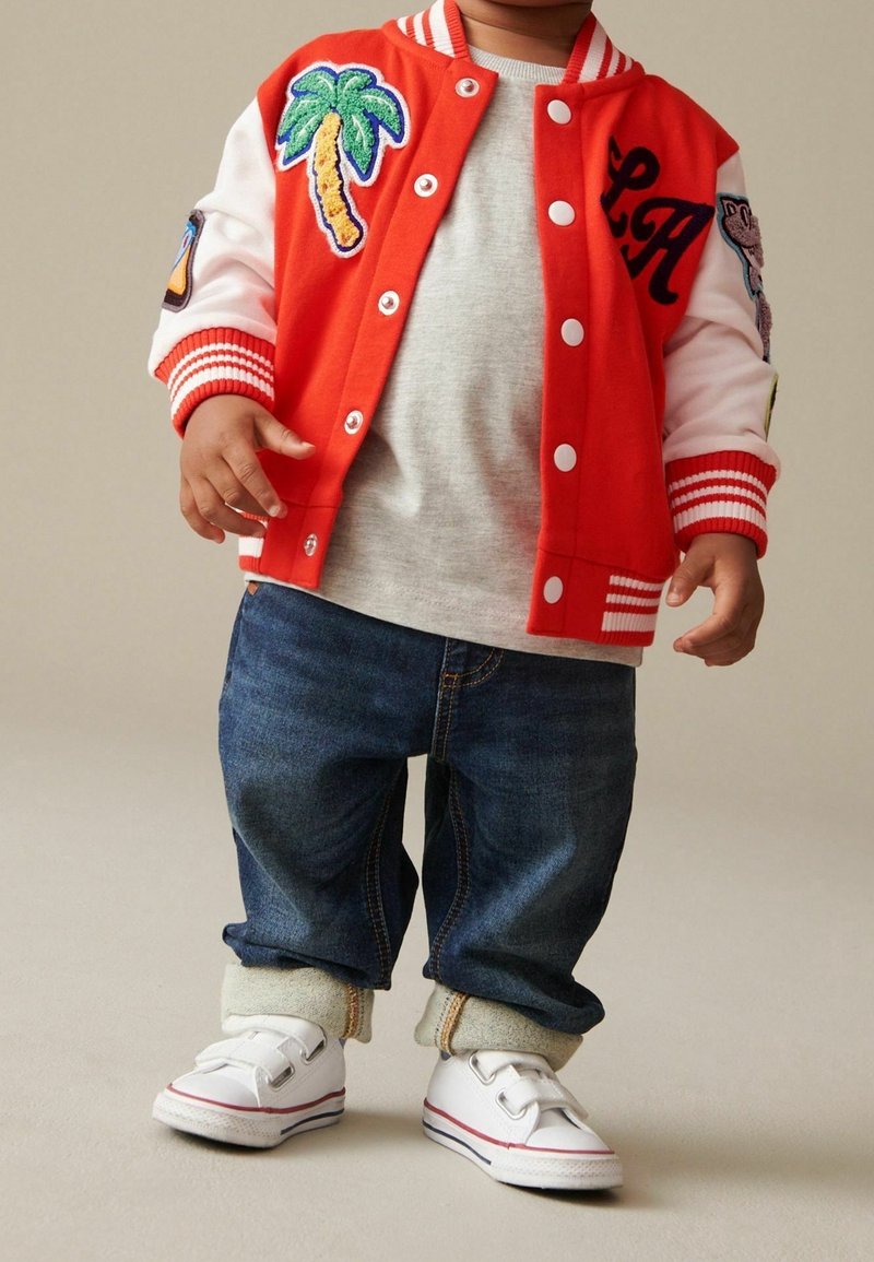 Tout-petit portant une veste rouge et blanche avec un écusson palmier, un t-shirt gris, un jean bleu avec revers et des baskets blanches à scratchs.