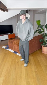Grijze zip-up hoodie en bijpassende broek, samen met een groene pet en zilveren sneakers. Gevoelige, comfortabele materialen in een casual pasvorm.