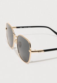 Kuldne metallraamiga päikeseprillid üle suurte ruudukujuliste läätsedega; mustad sangad "QUAY" brändinguga; hallid toonitud läätsed.