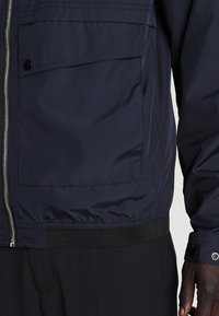 Navy blue jacket with a zip closure, elasticized hem, and chest pocket. Made of smooth material with a matte finish.