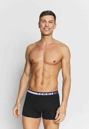 Czarne bawełniane bokserki z niebiesko-białym elastycznym pasem w rysunek "TOMMY HILFIGER", dopasowany krój. Model pozuje z uśmiechem.