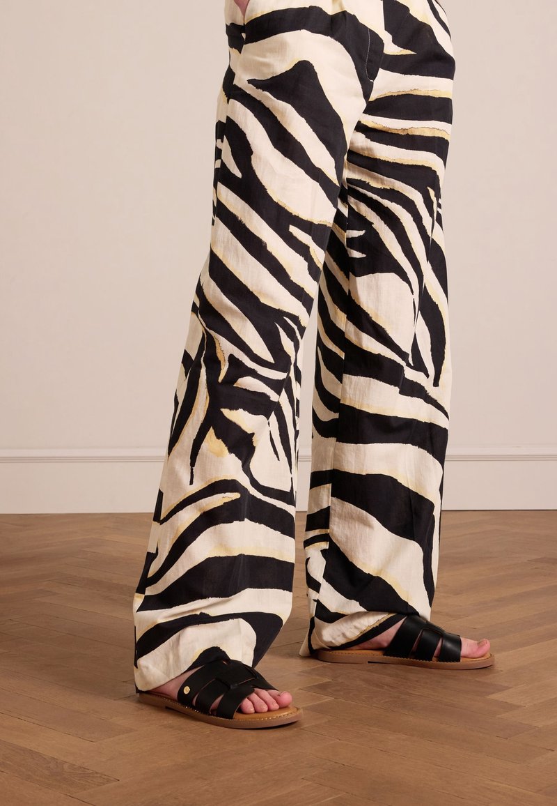 Personne portant un pantalon ample à rayures zèbre noir et blanc et des sandales plates noires, debout sur un sol en bois contre un mur clair.