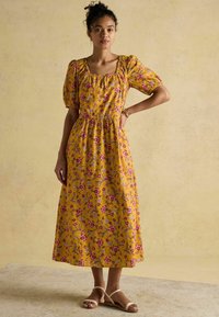 Kollane lilleline kleit lühikeste puhvärvidega, ruudu kaelusega ja elastse vöökohtadega. Omab roosa ja sinise lillemustriga. Kanda kergete sandaalidega.