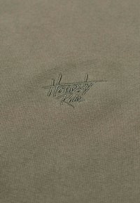 Närbild av olivgrön tyg med liten, broderad text "Homerity Rules" i matchande tråd centrerad på materialet.