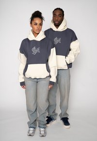 Oversized hoodies in marineblauw en crème, met een logo op de borst, gecombineerd met losse spijkerbroeken. Zachte stof met een gladde textuur.