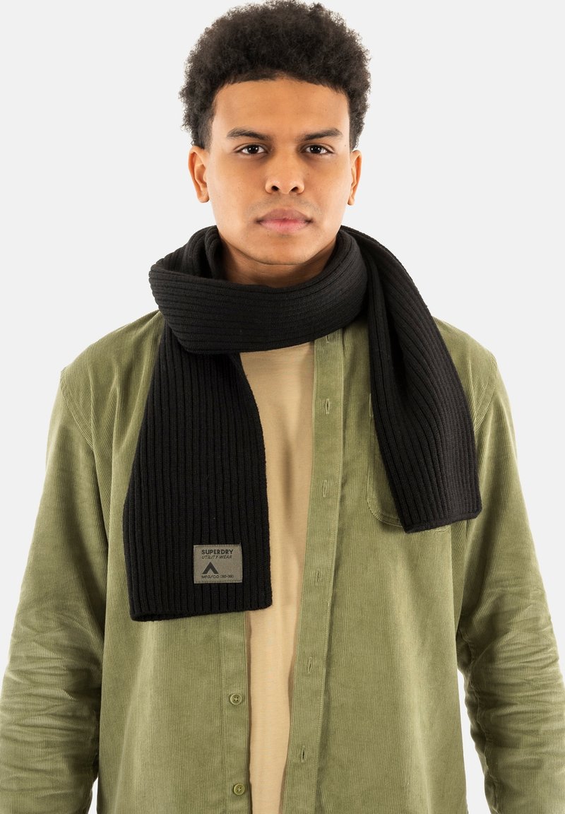 Sort ribbet tørklæde lavet af tekstureret stof, med et mærke, hængende om halsen, parret med en olivengrøn kordfløjlsskjorte.