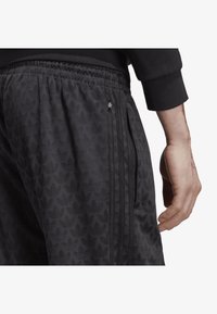 Svarta träningsbyxor med ett subtilt lövtryck, elastisk midja och fickor med dragkedja på sidorna. Försedda med detaljer i form av tre ränder.