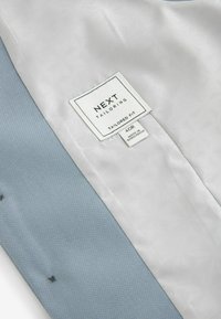 Blazer ajusté à l'intérieur avec doublure en satin gris clair présentant un motif géométrique subtil, accompagné d'un tissu texturé bleu. Étiquette d'entretien visible.