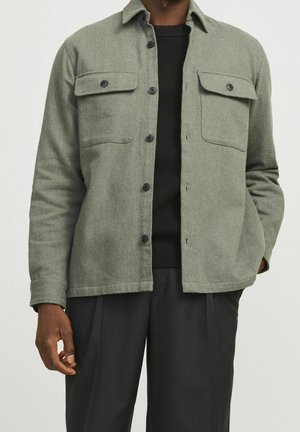 Homme portant une veste boutonnée vert olive sur une chemise noire et un pantalon noir, avec une main dans la poche.